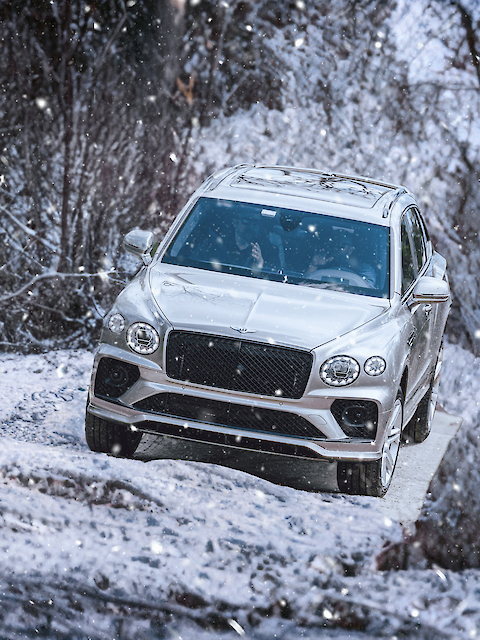 A silver SUV is driving through a snowy forest landscape with snow falling.