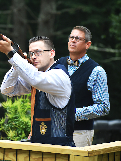 Two men are at a shooting range; one aims a shotgun while the other observes. They are wearing protective vests and glasses.