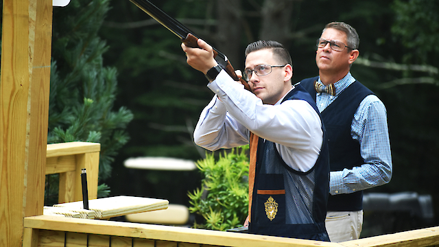 Two men are at a shooting range; one aims a shotgun while the other observes. They are wearing protective vests and glasses.