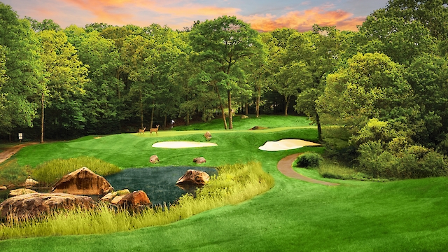 A picturesque golf course with lush greenery, a pond with rocks, and sand traps under a colorful sunset sky.