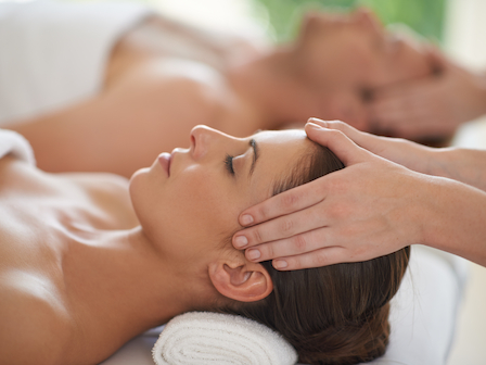 The image shows two people receiving head massages, lying down with towels under their heads, in a relaxing spa setting.