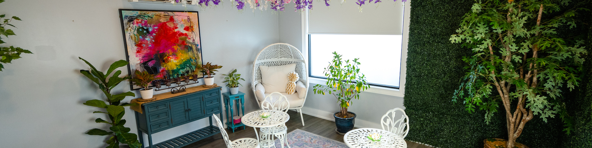 The image shows a cozy room with white furniture, vibrant plants, and colorful flowers hanging from the ceiling, creating a serene atmosphere.