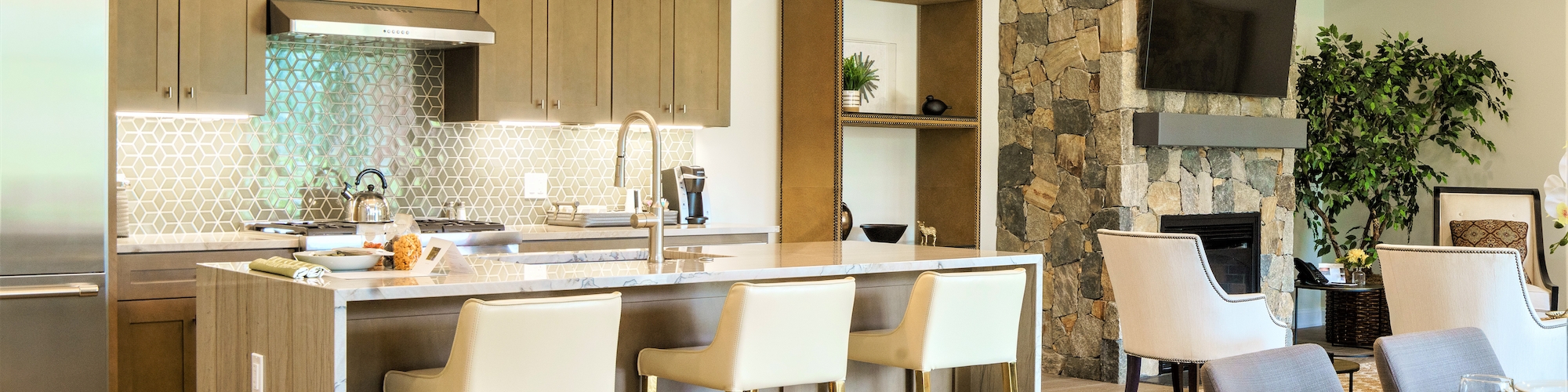 Modern kitchen with an island, barstools, and open living area featuring a fireplace and TV. Neutral tones and wood accents complete the look.