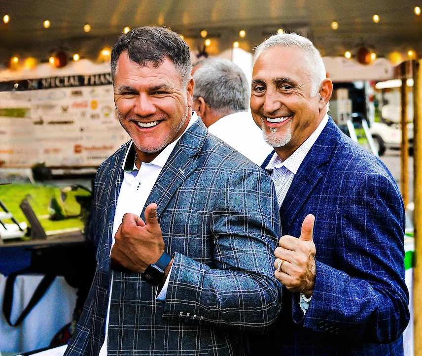 Two men in suits giving thumbs up, smiling outdoors at an event.