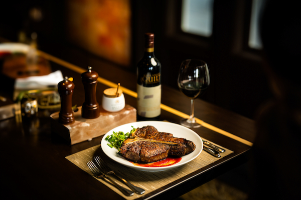 A plate with steak and greens, a bottle of red wine, glass, salt and pepper shakers, and cutlery on a dining table.