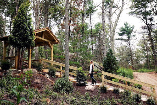 A person walks along a wooden pathway amid trees and greenery in a forested area with a sloped landscape and small pavilion.