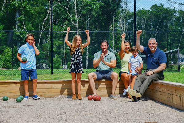 A group of people, including children and adults, are sitting and standing around a bocce court with raised arms, celebrating outdoors.