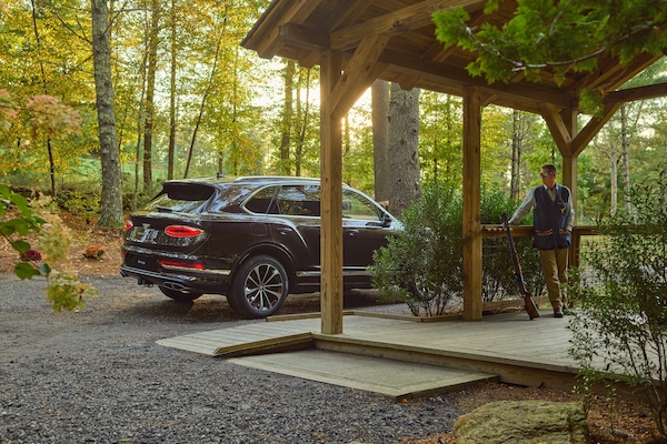 A person stands on a wooden porch beside a dark SUV in a wooded outdoor setting with autumn foliage and flowers.