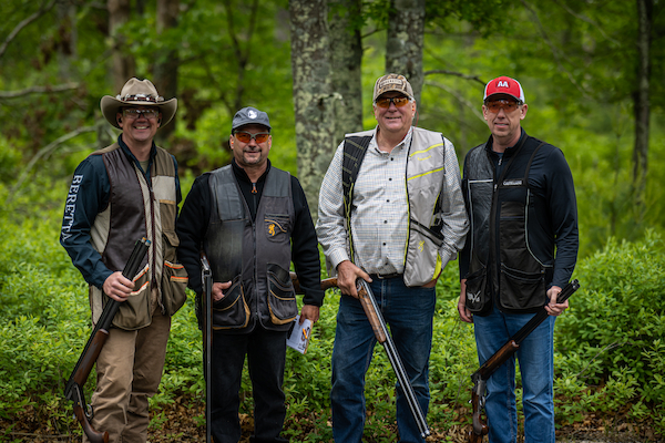 Four people wearing casual and shooting gear, holding rifles, standing in a wooded outdoor area, posing for the photo.