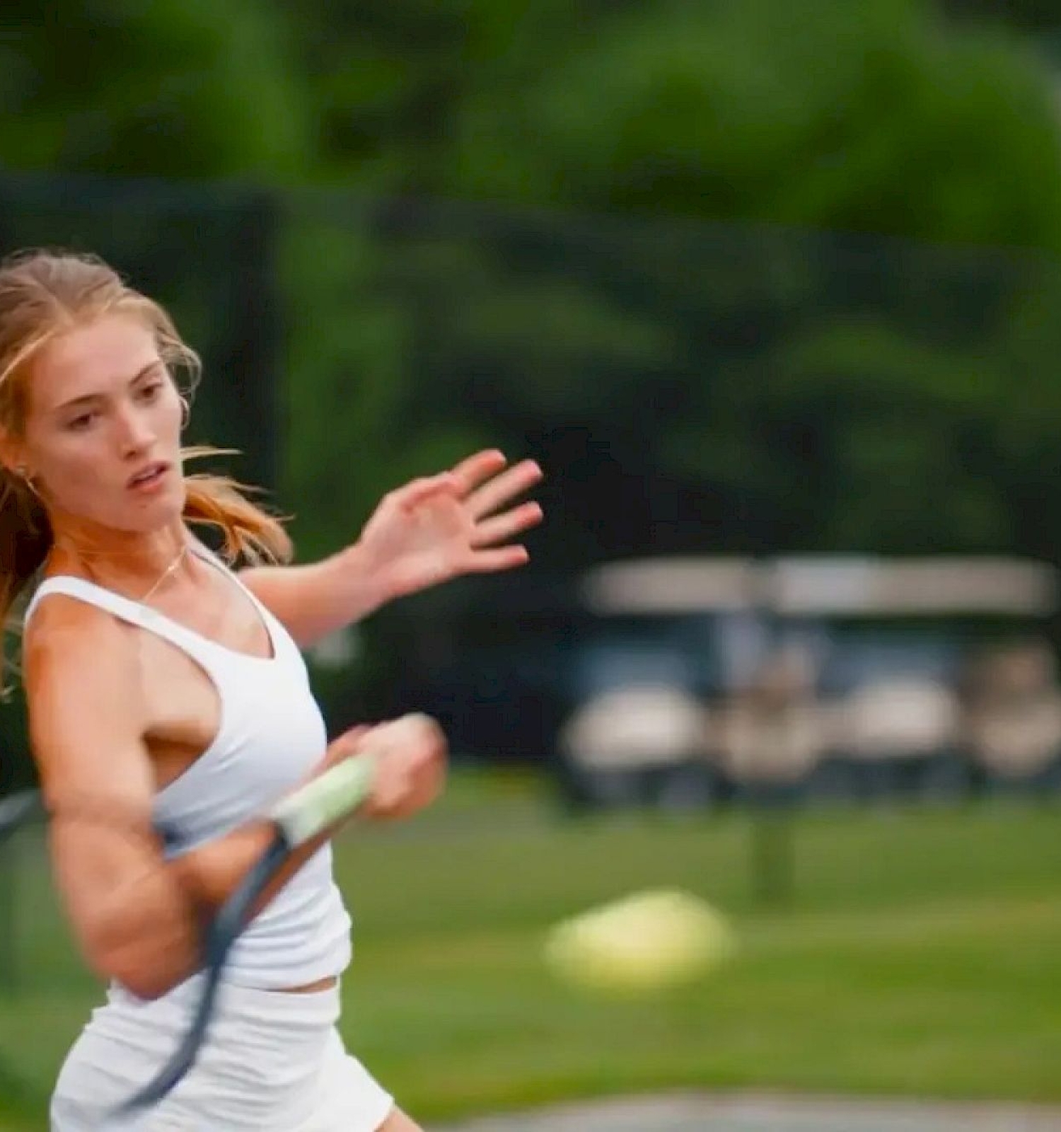 A person is playing tennis, swinging a racket to hit a tennis ball on a green outdoor court with blurred trees in the background.