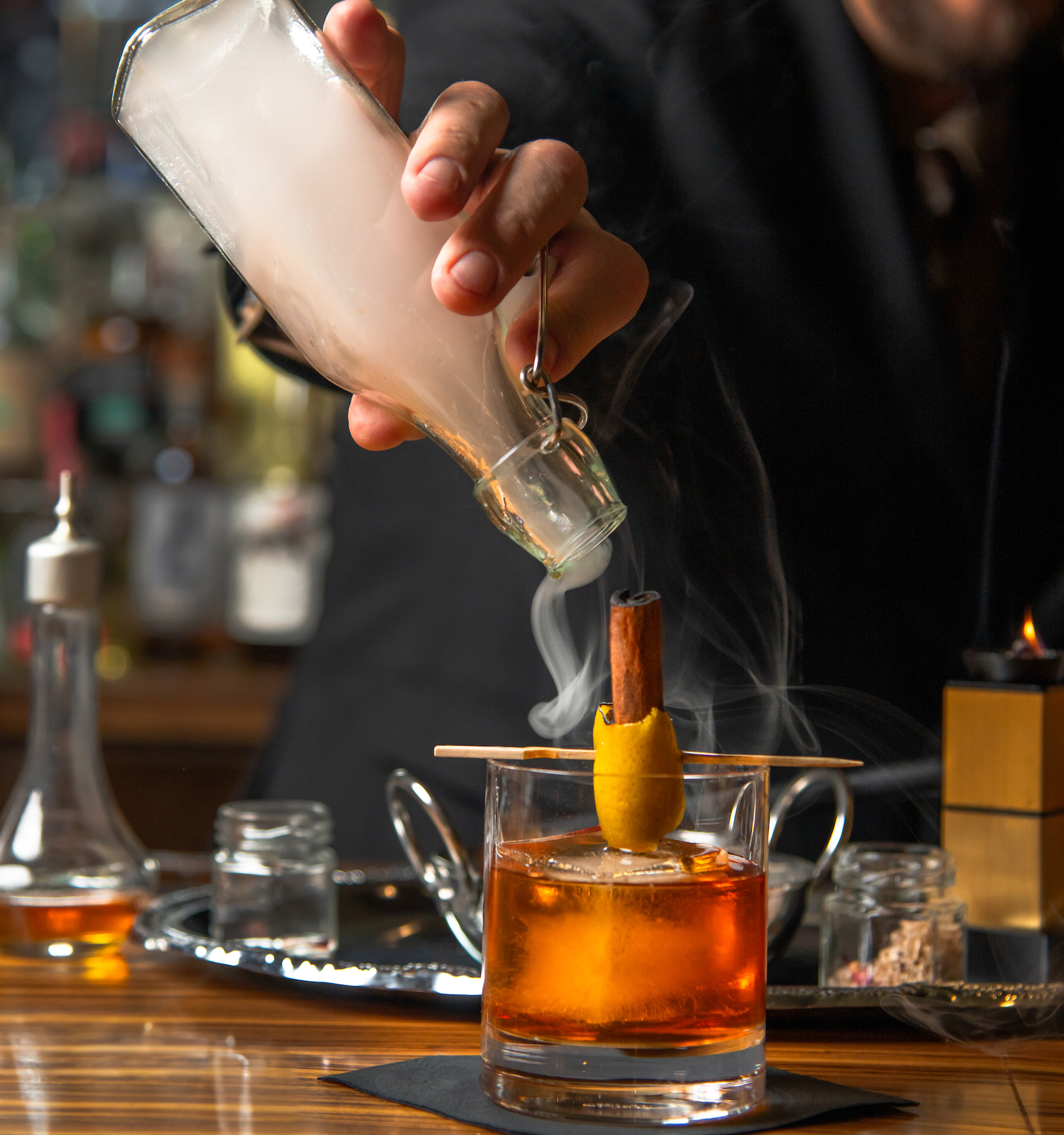 A bartender adds smoke to a cocktail with a cinnamon stick and peel garnish, creating a dramatic presentation.