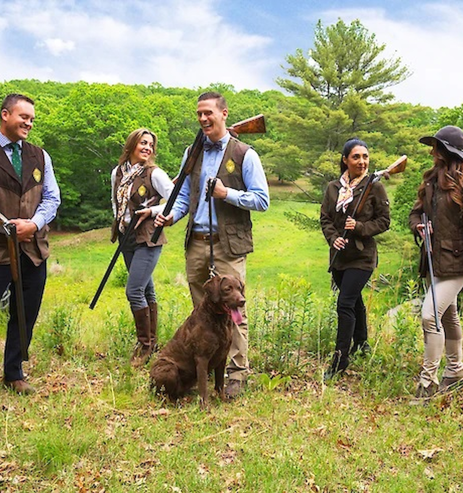 A group of people with rifles and a dog stand outdoors in a field, surrounded by greenery, under a partly cloudy sky.