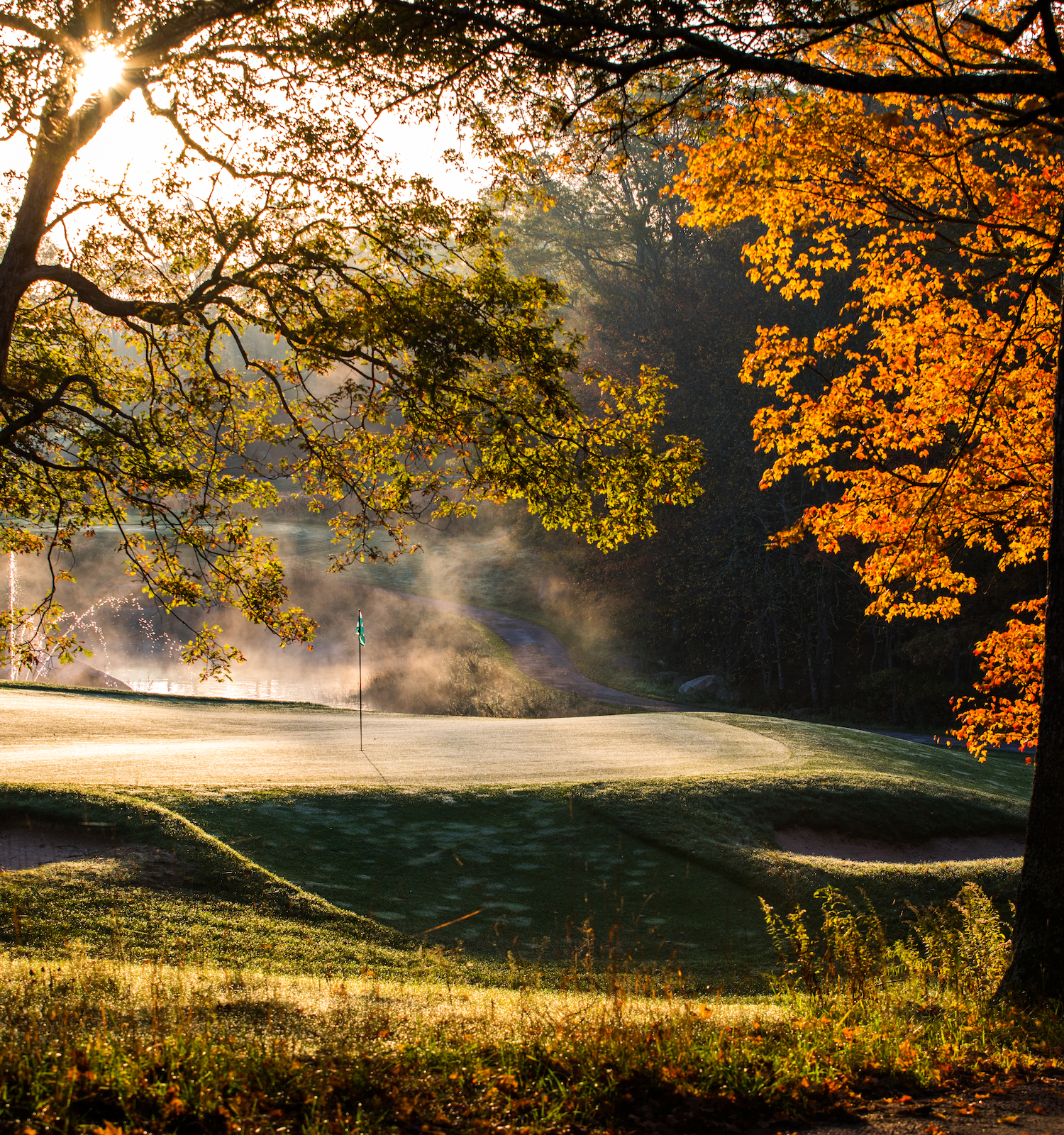 Autumn scene with a sunlit golf course surrounded by trees showing vibrant fall foliage. Mist lightly hovers over the grass.