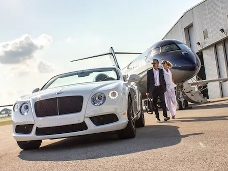 A white luxury car parked on a tarmac, a private jet in the background, and two people walking towards it from the aircraft.