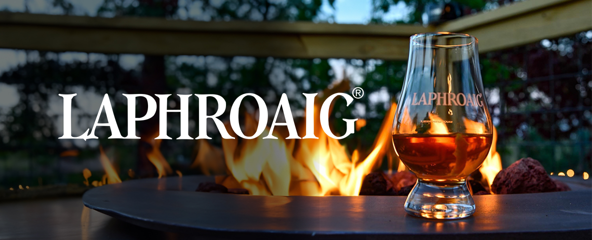 A glass of Laphroaig whisky sits on a table with a fire in the background, creating a warm and inviting atmosphere.