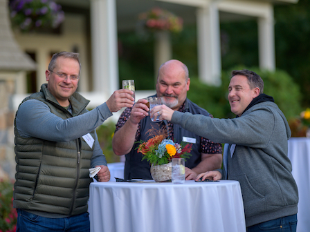 Three people are toasting drinks at a table outdoors, with flowers in the background.