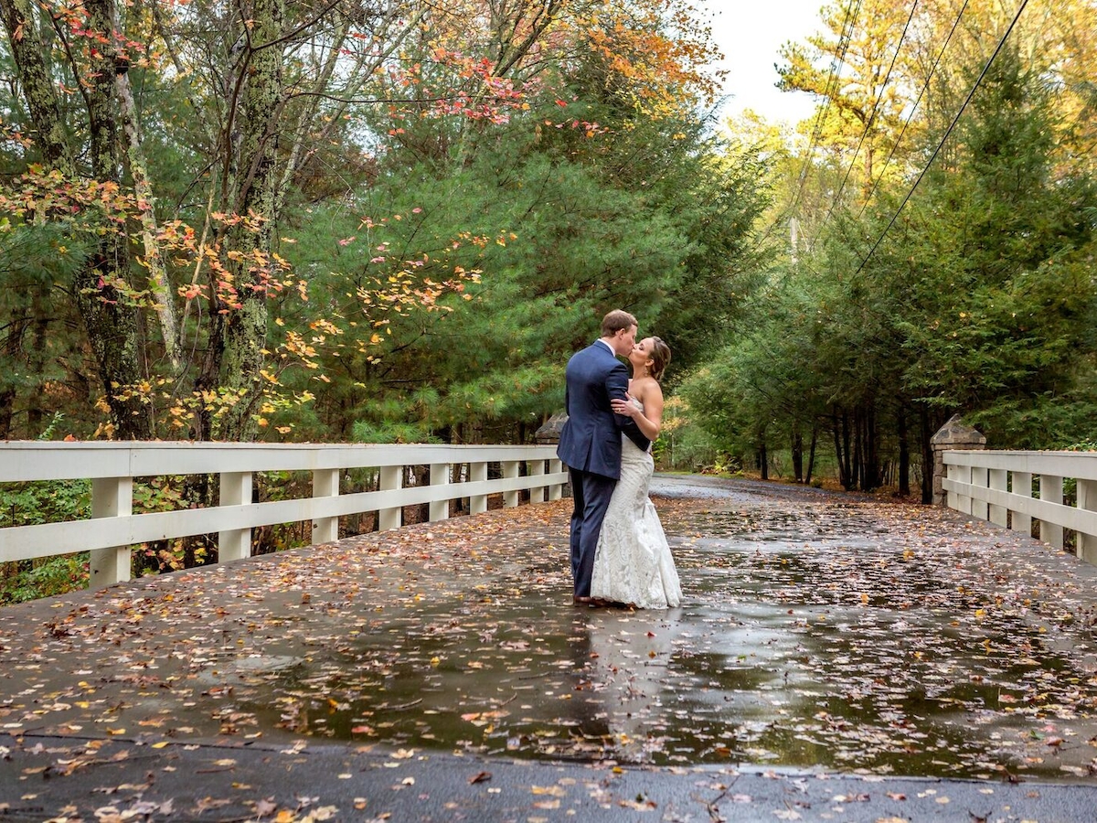 A couple embraces on a leaf-strewn bridge surrounded by trees during autumn, creating a romantic and picturesque scene.