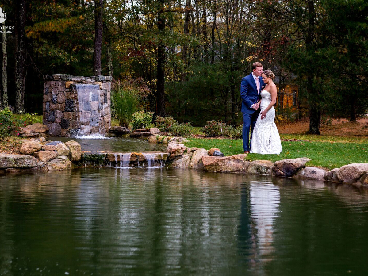 A couple stands by a pond in a wooded area, dressed formally. A small waterfall and stone structure are in the background.