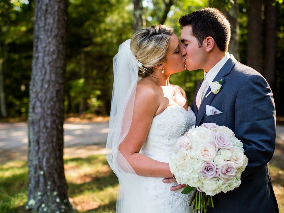 A couple is kissing outdoors, with the bride in a white dress holding a bouquet, and the groom in a suit. They're standing by trees.