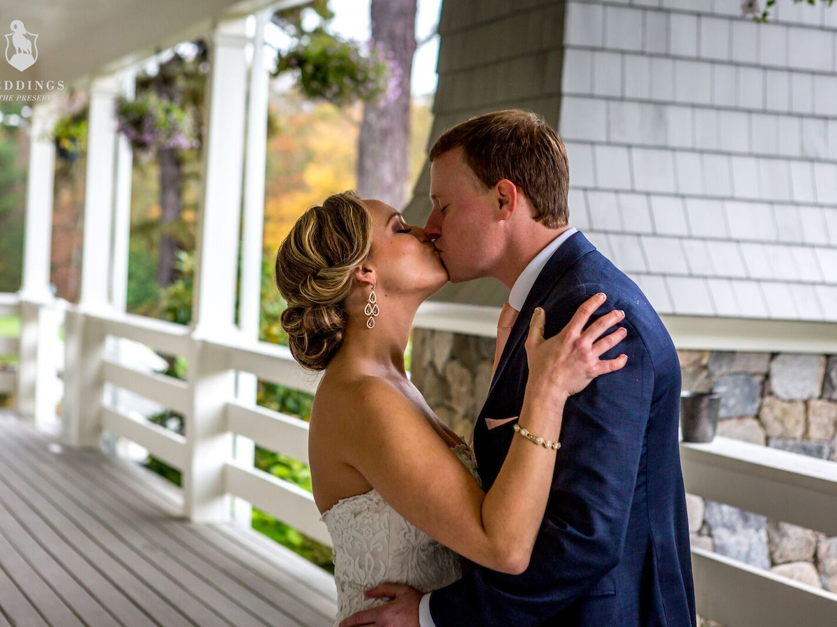 A couple is kissing on a porch, possibly during a wedding, with trees and a stone structure in the background.