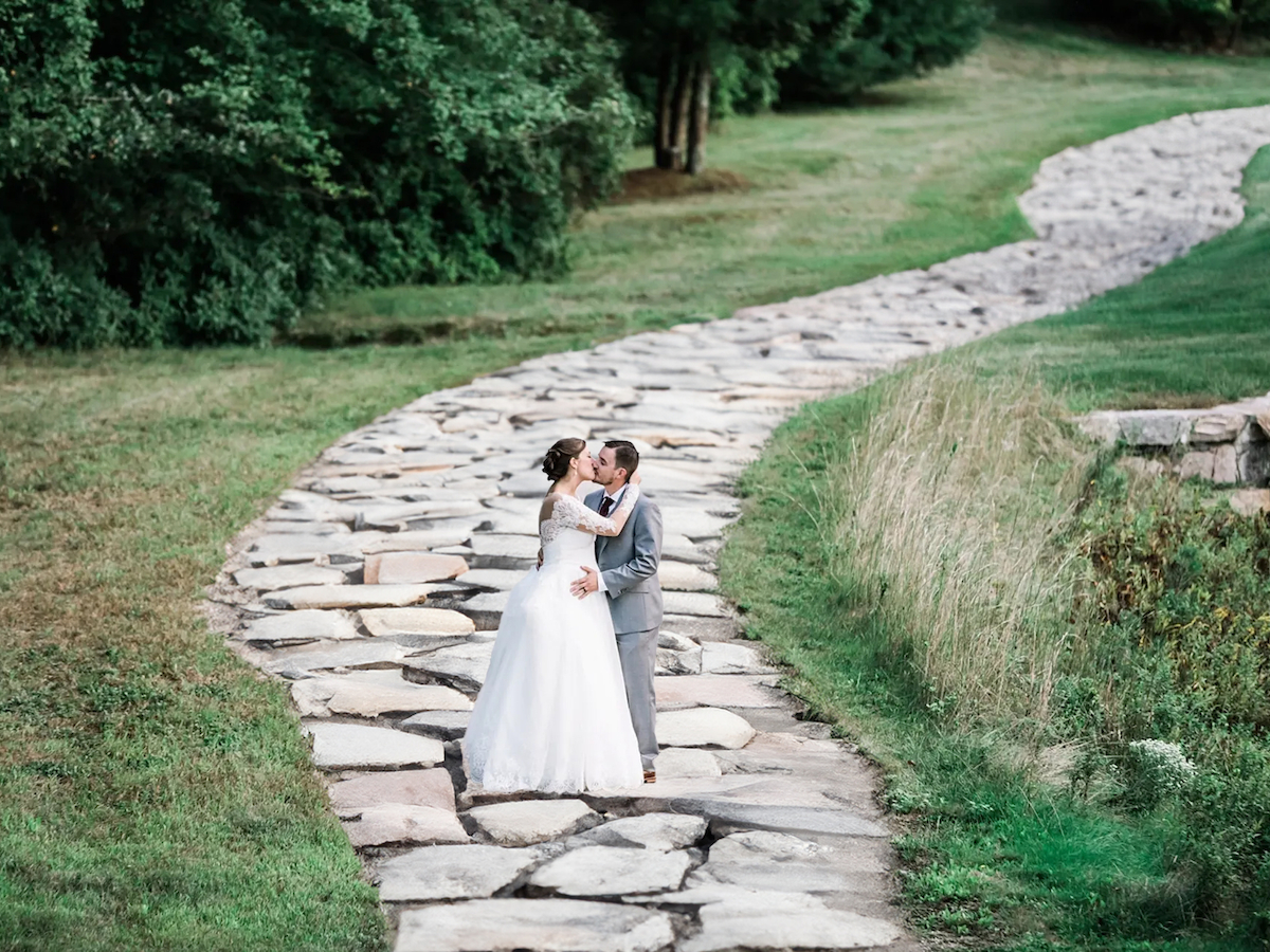 A bride and groom stand on a stone path, surrounded by greenery, sharing a romantic moment.