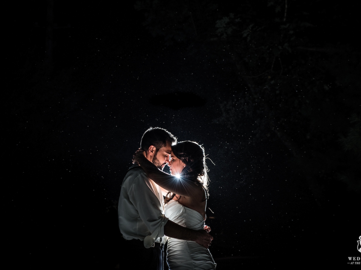 A couple is embracing in the rain at night, backlit to create a romantic silhouette.