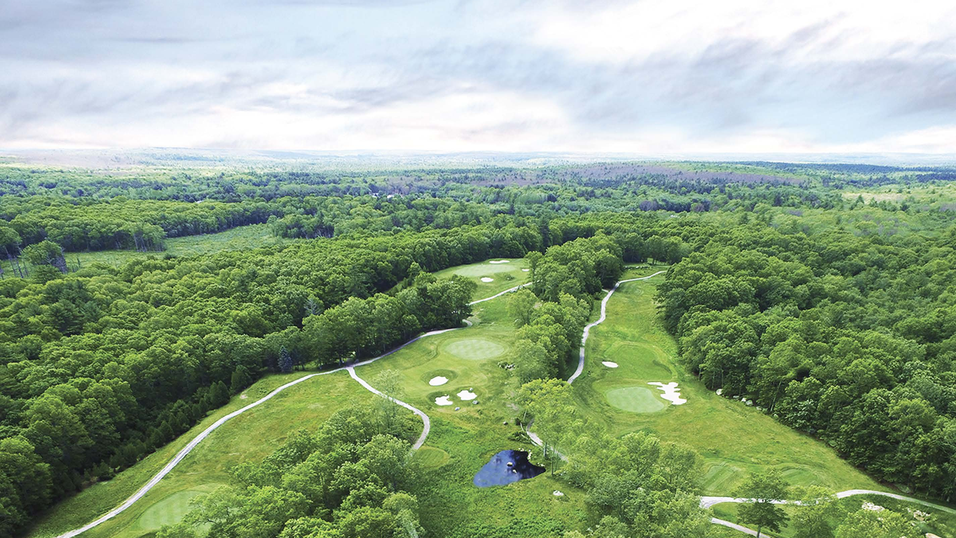 An aerial view of a green, sprawling landscape featuring a golf course with pathways, surrounded by dense forest under a cloudy sky.