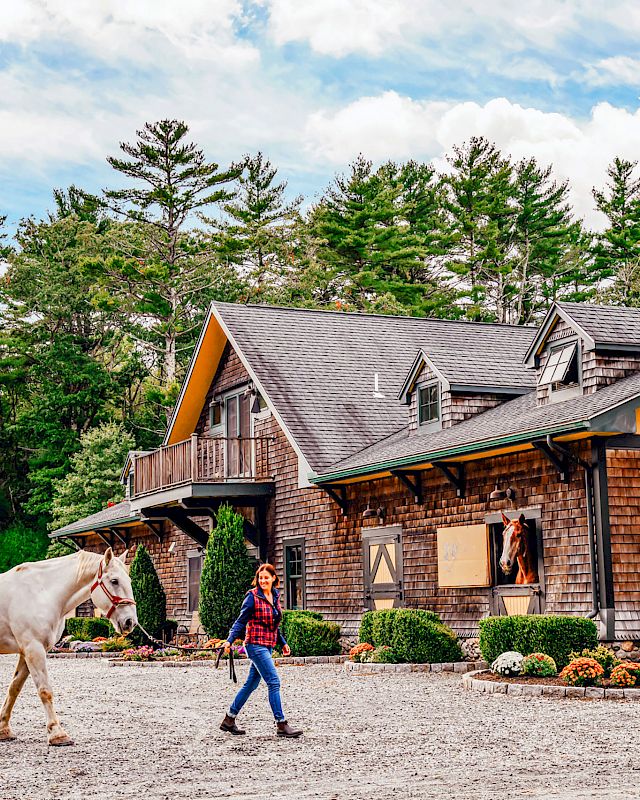 A person is leading a white horse near a large barn surrounded by trees and shrubs, with a clear sky overhead.