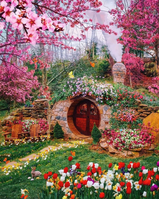 A whimsical garden scene features a round wooden door set into a hillside, surrounded by vibrant flowers and pink-blossomed trees.