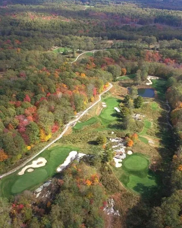 Aerial view of a golf course in a forested area with autumn foliage, featuring greens, sand traps, water hazards, and surrounding trees.