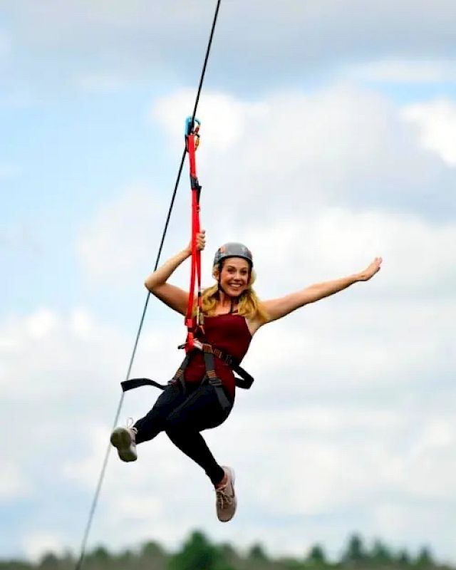 A person is zip-lining, wearing safety equipment and a helmet, with one arm outstretched, and smiling against a backdrop of clouds and trees.