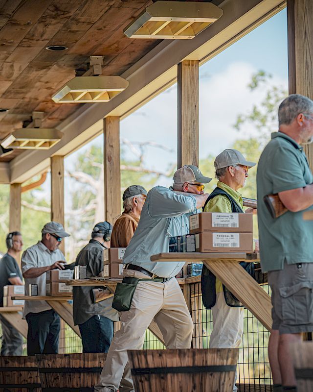 People are standing in a line at wooden booths, some wearing hats and vests, appearing to be engaged in an outdoor shooting or archery activity.