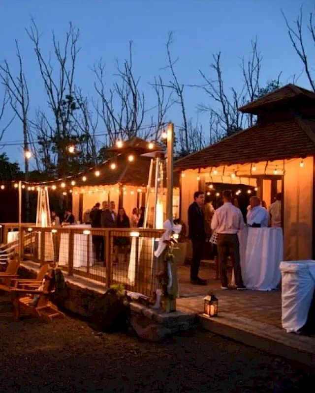 A warmly lit outdoor gathering with string lights, people mingling, and a welcoming wooden structure in the evening.