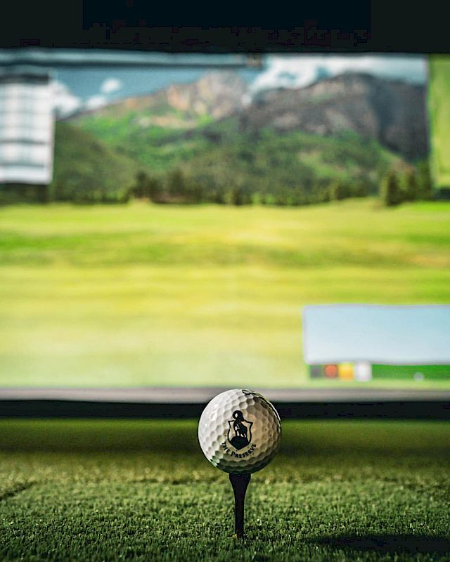 A golf ball on a tee in an indoor setup, with a large screen displaying a golf course in the background, creating a virtual golf experience.