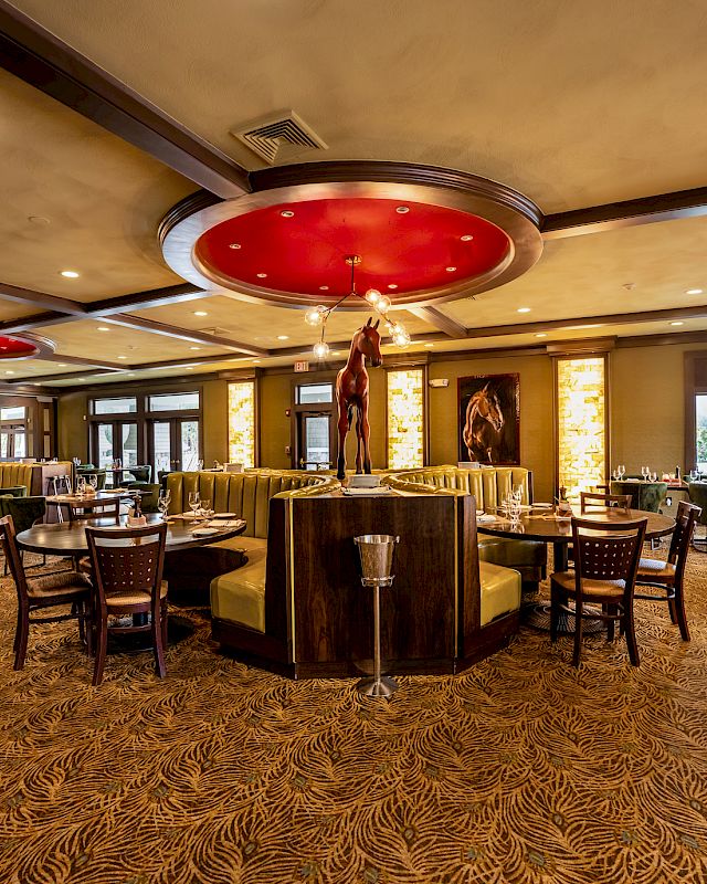 A spacious, well-lit restaurant interior featuring round tables, booths, decorative lighting, and artistic statues. The decor is warm and inviting.