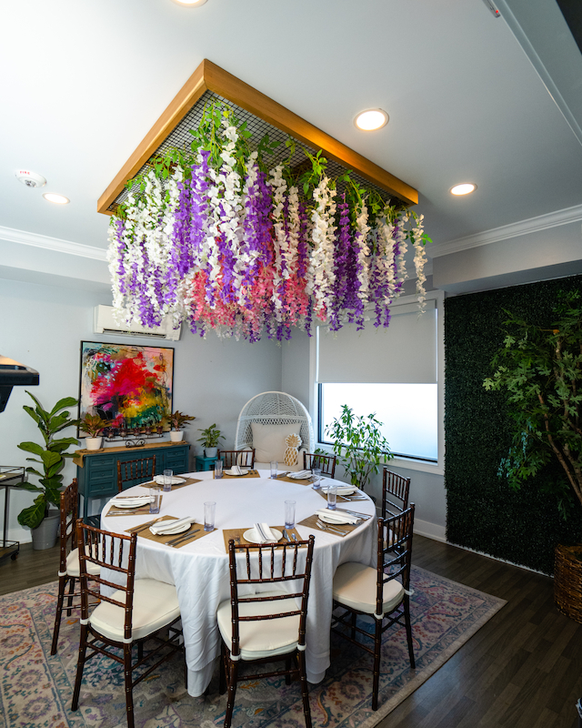 A dining area with a round table, chairs, and a floral ceiling decoration, set in a room with plants and artwork.