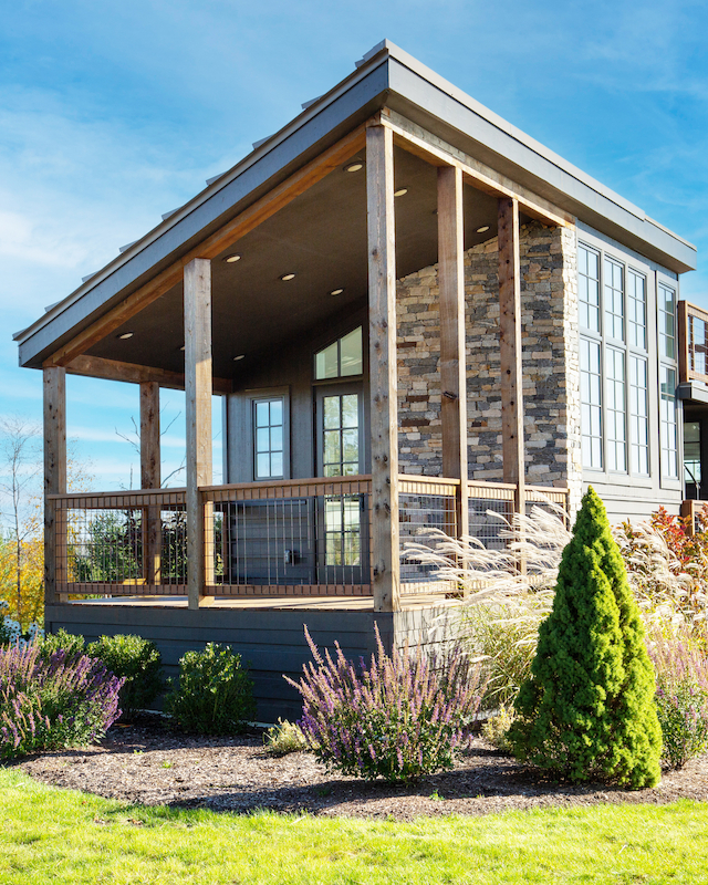 A modern house with large windows and a wooden porch, surrounded by a neatly landscaped garden on a sunny day.