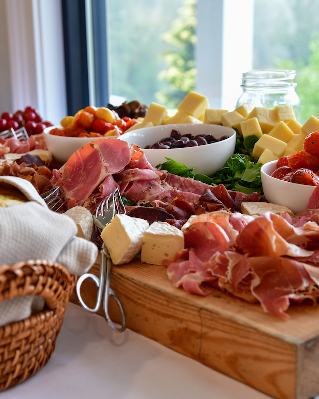 A charcuterie board with prosciutto, cheese cubes, grapes, olives, and bread in a basket, set on a table by a window.