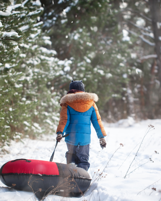 A person in winter clothes pulls a snow tube through a snowy forest path, surrounded by trees, under a gentle snowfall.