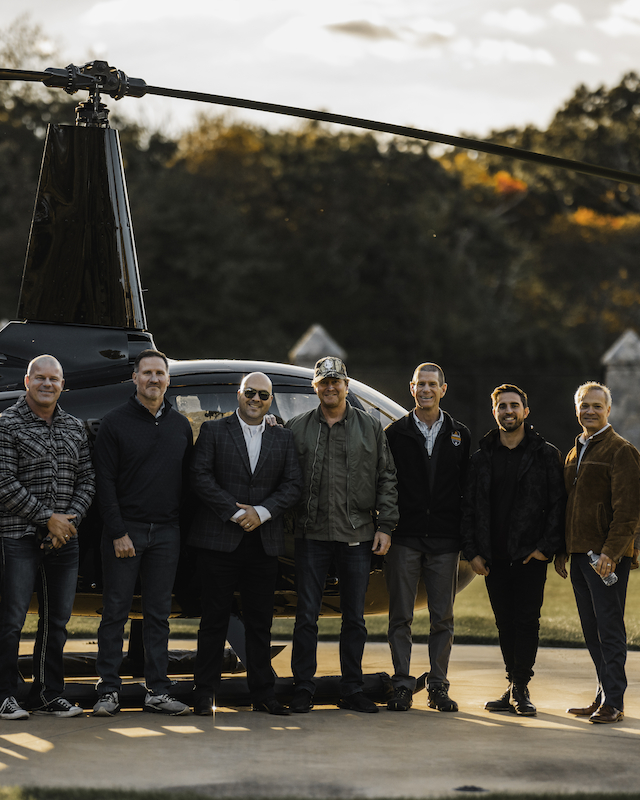 A group of nine people standing in front of a helicopter on a paved surface with trees in the background.