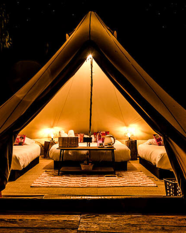 A cozy, illuminated glamping tent with two beds, a rug, and lamps, set up in a rich nighttime setting, creating a warm ambiance.