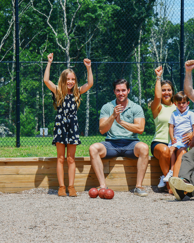 A group of people, including kids, celebrate on a bocce court, raising their arms. Trees and a fence are visible in the background.