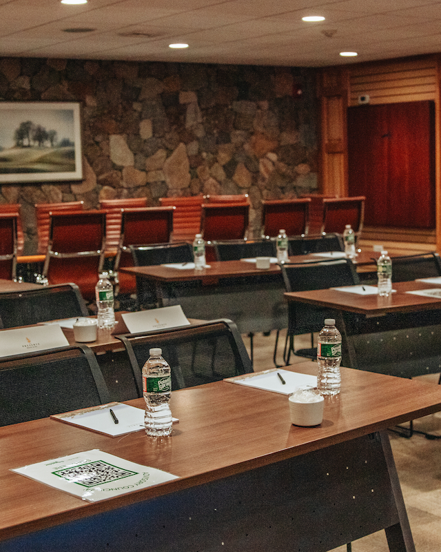 A conference room with tables, chairs, bottled water, and notepads set up for a meeting, featuring a stone wall and framed picture.