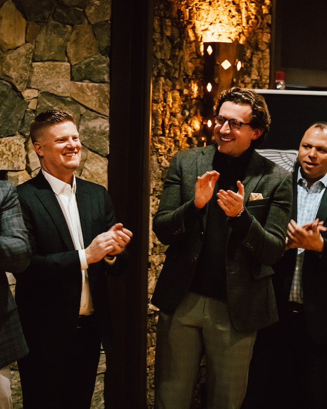 A group of four people in formal attire clapping, standing in a warmly lit setting with stone walls and a banner partially visible.