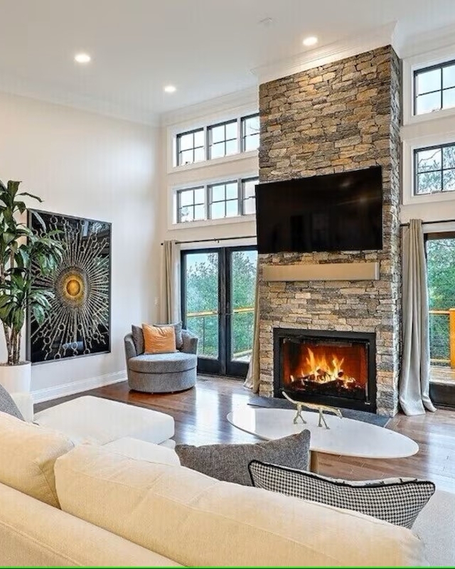 A cozy living room with a stone fireplace, large windows, TV, modern furniture, and warm lighting, creating a welcoming atmosphere.