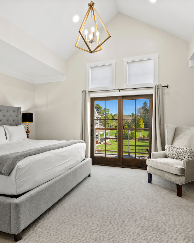 A spacious bedroom with a large bed, armchair, chandelier, and French doors leading to a deck with garden views.