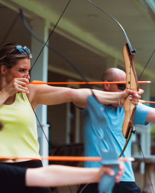 A group of people practicing archery outside, each aiming bows with arrows, standing in line and focusing on their targets.