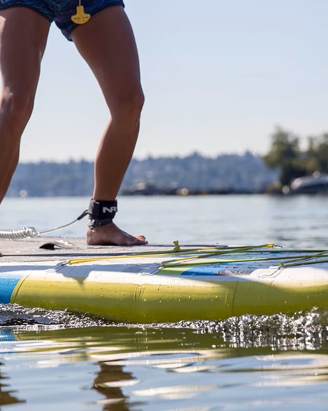 A person is stand-up paddleboarding on a calm body of water, using a paddle and wearing a leash on their ankle.