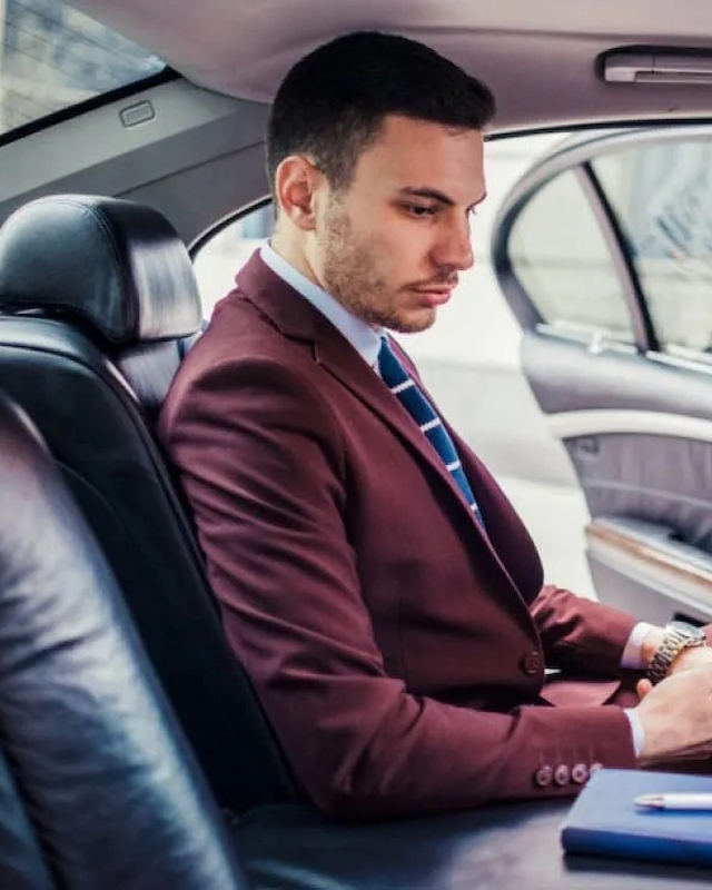 A man in a maroon suit is seated in the back of a car, looking at his phone, with a notebook beside him on the seat.