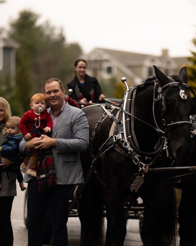 A family poses beside a horse-drawn carriage, with a driver ready to steer the two black horses in front of a suburban background.